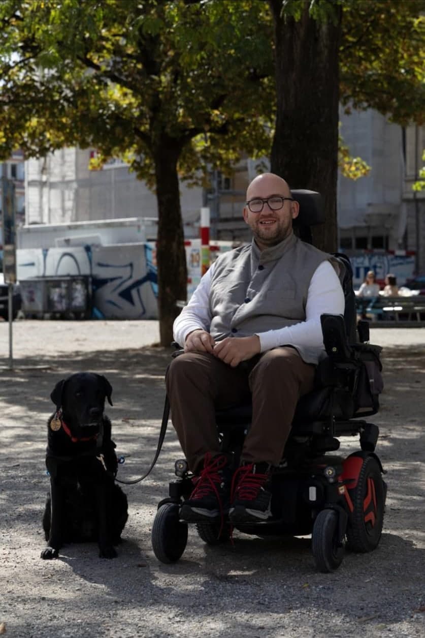 Matyas Sagi-Kiss sitzt lächelnd in seinem Elektrorollstuhl auf einem sonnigen Platz unter Bäumen. Links neben ihm sitzt sein schwarzer Assistenzhund an der Leine.